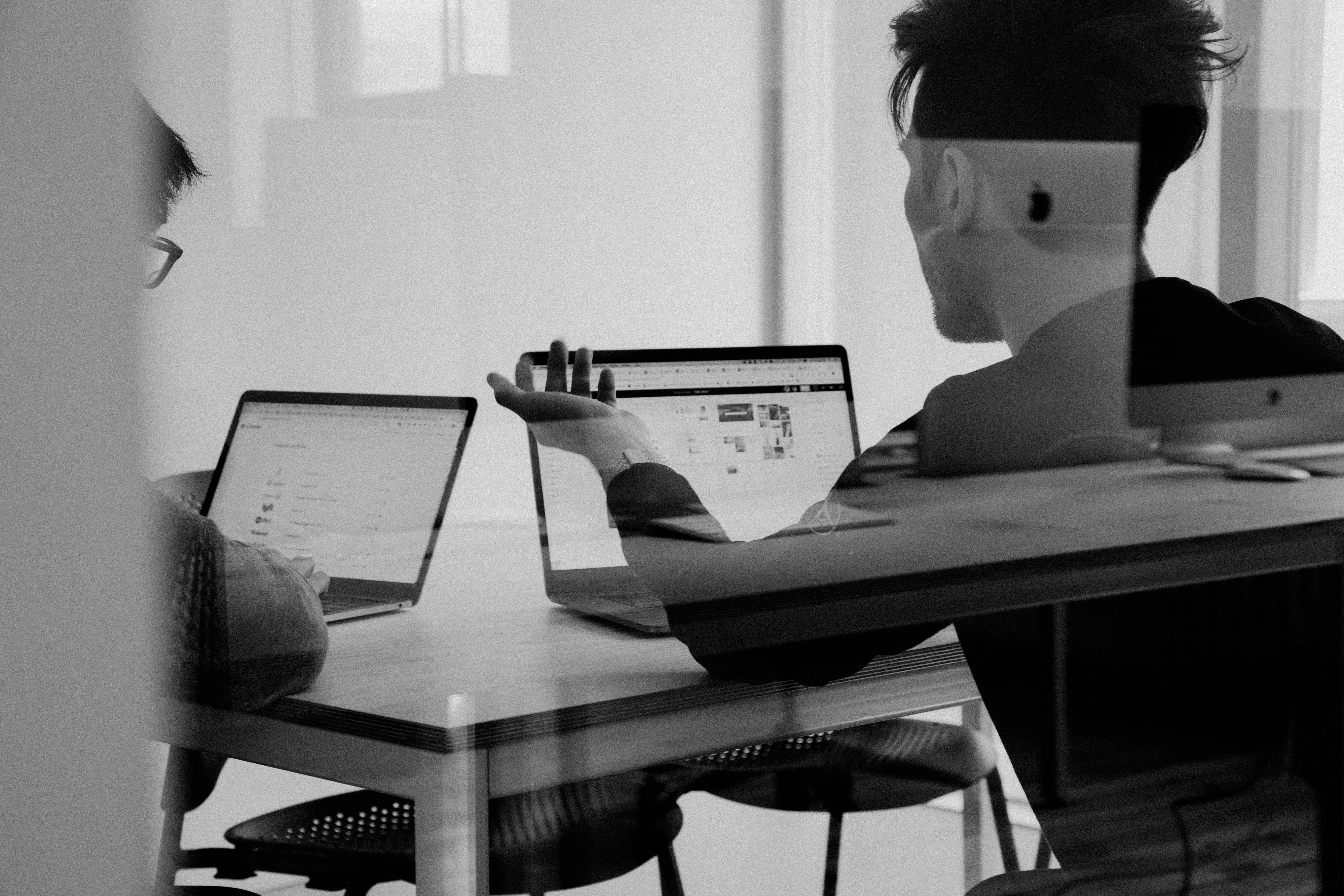 Zwei Personen arbeiten gemeinsam an Laptops an einem Schreibtisch, Schwarz-Weiß-Aufnahme